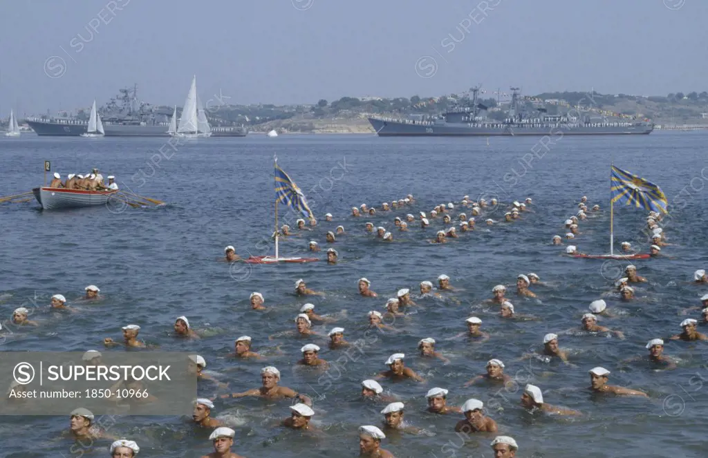 Russia, Sevastopol, Navy Day. Sailors Swimming In The Sea In Formation With Battleships Behind.