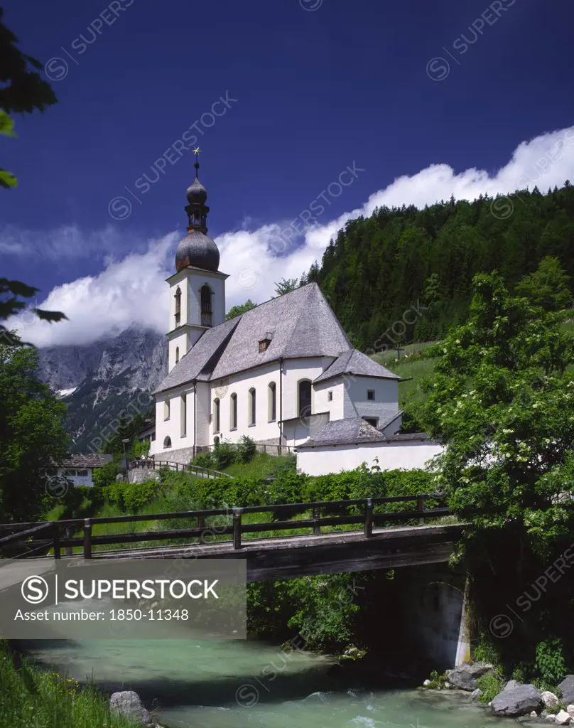 Germany, Bavaria, Ramsau, View Over River And Wooden Bridge Towards Ramsau Church And Wooded Hillside Beyond.