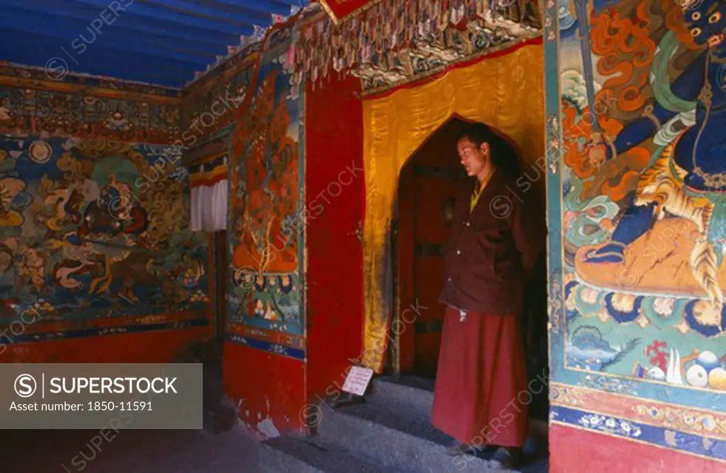 Tibet, Sakya, Monk Standing In A Doorway With Brightly Painted Religious Scenes And Deities Around Him.
