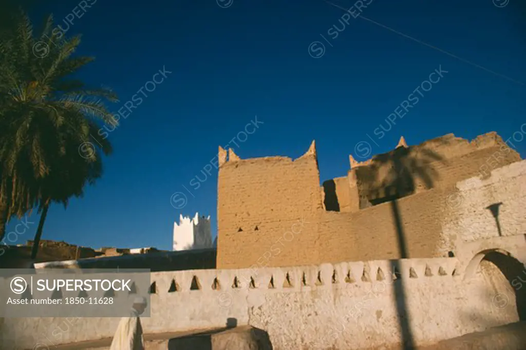 Libya, Ghadames Oasis, Man Walking Past Whitewashed Wall With Mud Brick Town Walls And White Mosque Minaret Behind