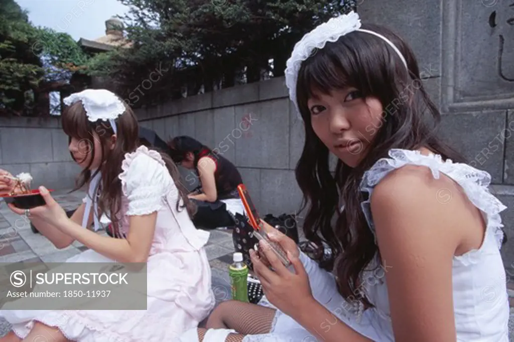 Japan, Honshu, Tokyo, Harajuku District. Two Teenage Girls Sitting Together With One Eating And The Other Using A Mobile Phone