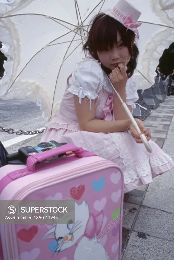 Japan, Honshu, Tokyo, Harajuku District. Portrait Of Young Teenage Girl Wearing A Pink And White Dress Holding A Parasol With A Pink Suitcase In The Foreground