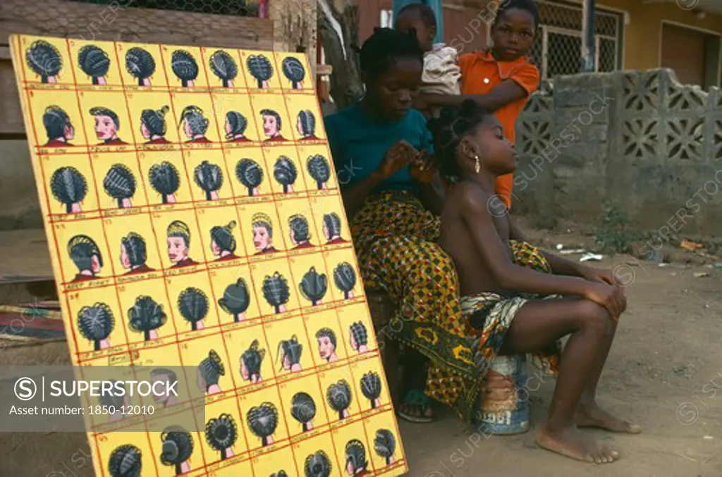 Nigeria, Lagos, Hairdresser Braiding Hair Of Young Girl At Roadside Beside Board Displaying Different Hairstyles.