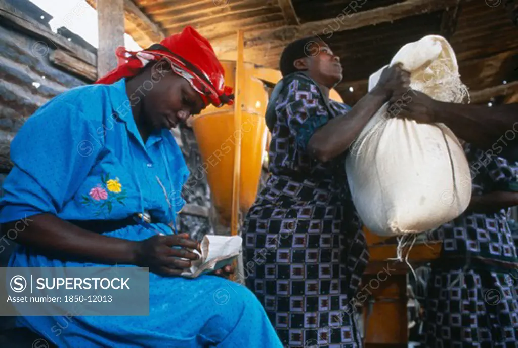Tanzania, Food Aid, Women Lifting Up A Sack Of Food Aid Next To A Woman Sat Writing On Piece Of Paper.