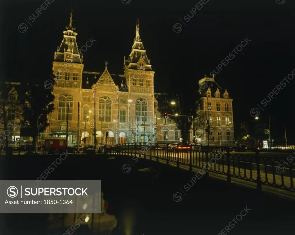 Holland, North, Amsterdam, Rijksmuseum Illuminated At Night Beyond A Bridge Over A Canal Reflected In The Water
