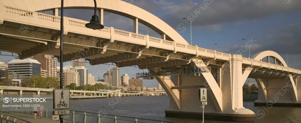 Australia, Queensland, Brisbane, Walkers Strolling Along The River Walk Beneath A Road Bridge Over The Brisbane River.