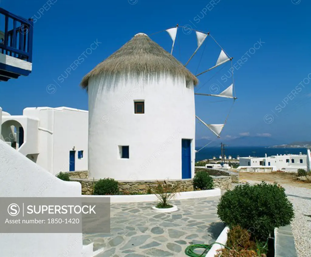 Greece, Cyclades Islands, Mykonos, 'White Windmill With Thatched Roof, Sails Have Flags At Tips, Blue Door, White Town Buildings & Coast Beyond. Nr.06.053579.00 '