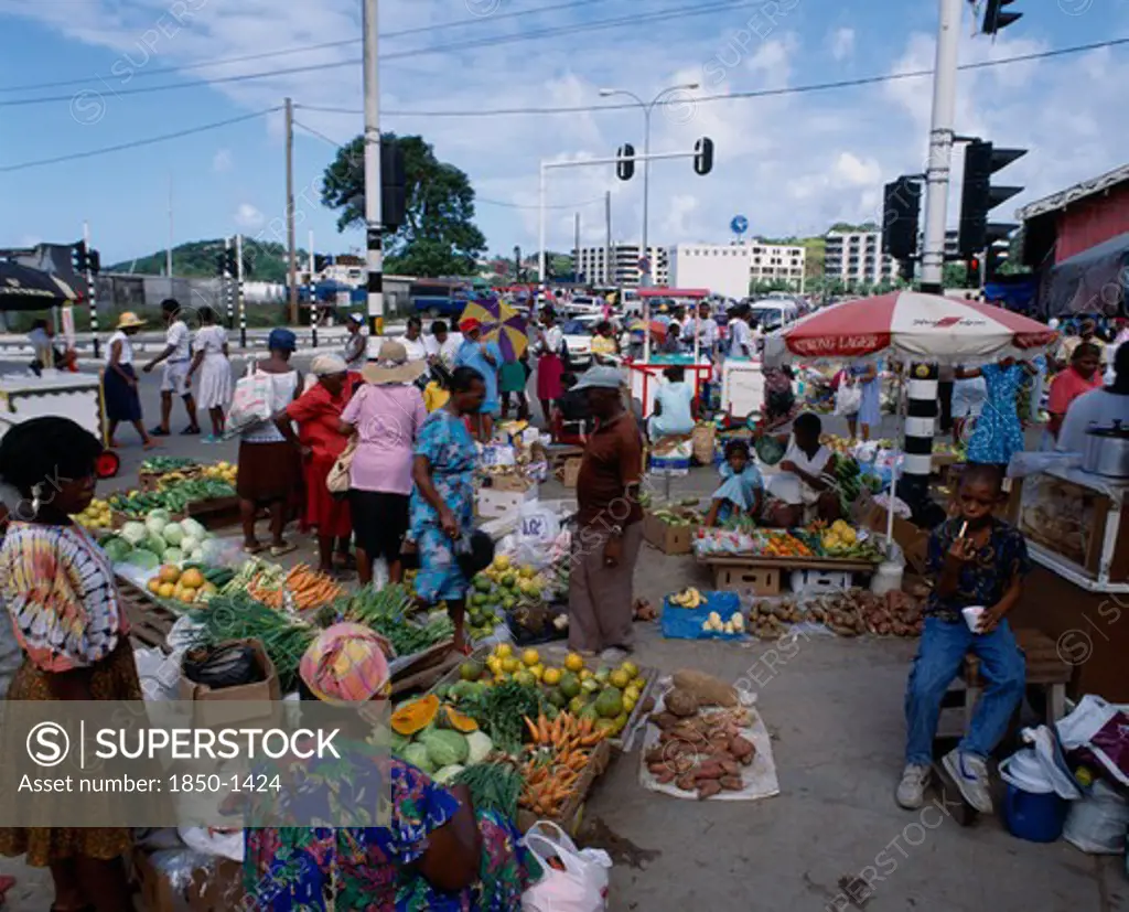 West Indies, St Lucia, Castries, 'Fruit And  Vegetable Street Market. Pumpkins, Carrots, Cabbage, Yams, Spring Onions, Bananas, Oranges Near Street With Traffic Lights And Ice-Cream Vendor In Background. '