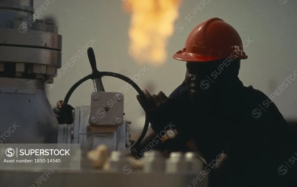 Nigeria, Industry, Portrait Of Oil Worker With Flare Of Burnt Off Gas Behind Him.