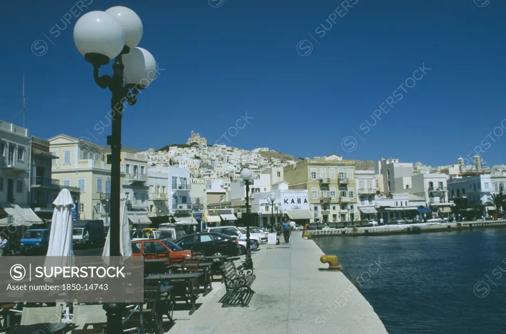 Greece, Cyclades Islands, Syros, Ermoupolis. The Town With Benches And Cafes At The Waters Edge With The Orthodox Church Anastasi On Top Of The Hill Bedind.