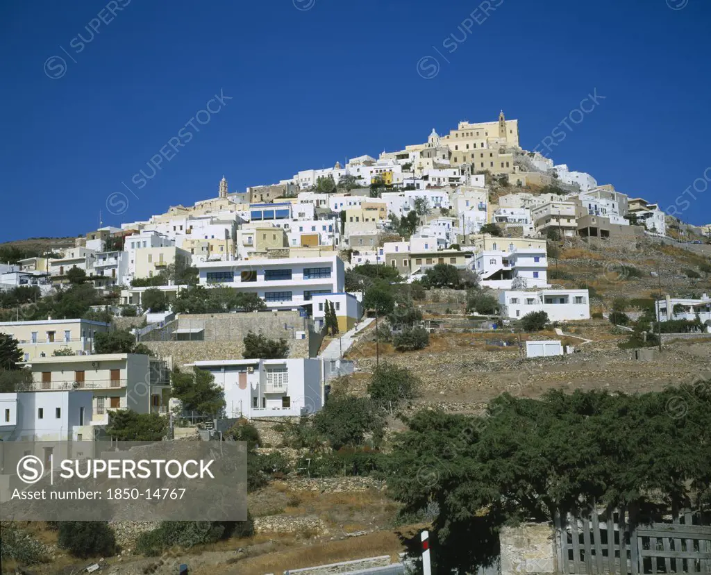 Greece, Cyclades Islands, Syros, Ermoupolis. The Catholic Quater Of Ano Syros And Church Of Ag. Yiorgios. Houses Covering Mountain Side With Trees And A Wall In The Foreground.