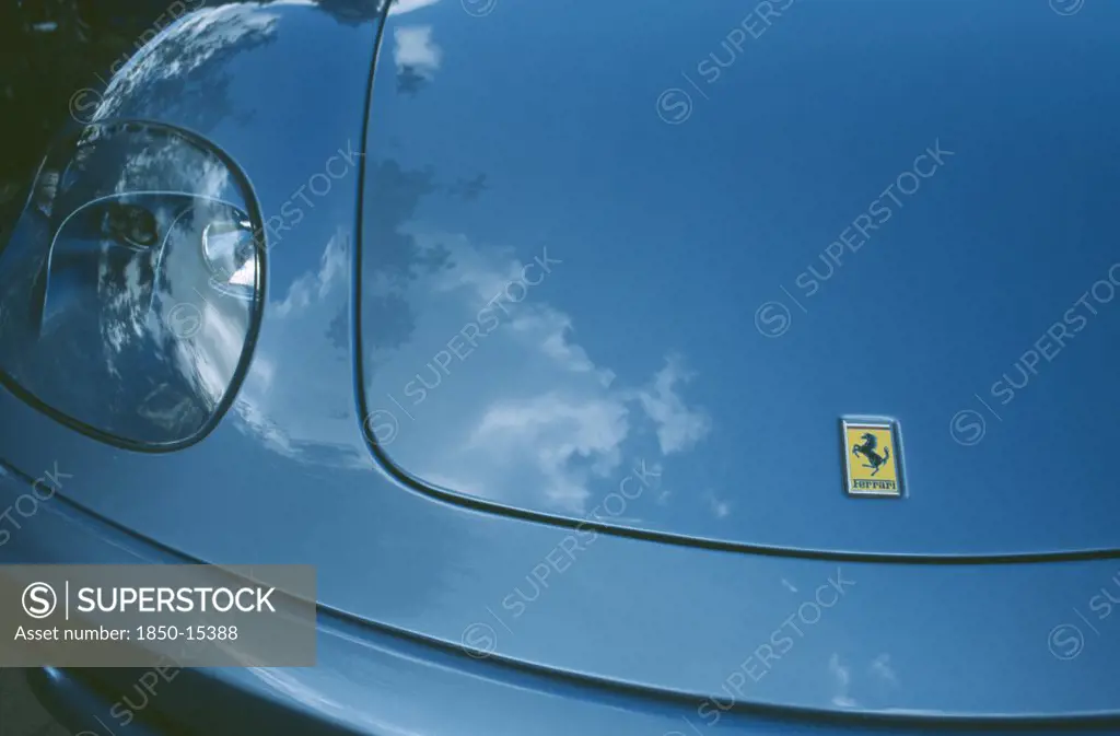 Transport, Road, Cars, Detail Of Blue Ferrari Emblem And Front Light With Clouds Reflected In Paintwork.