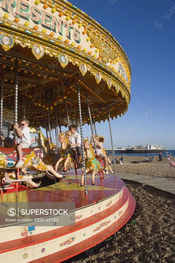 England, East Sussex, Brighton, Children Riding On A Fairground Carrousel On The Beach With Brighton Pier In The Distance