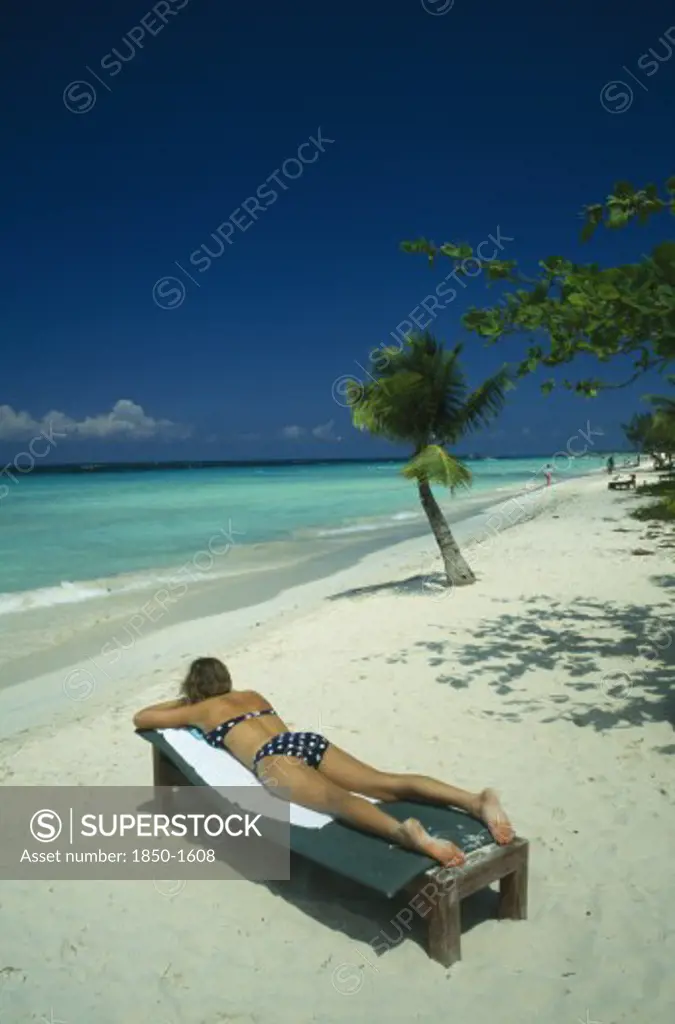 West Indies, Jamaica, Negril, Woman Sunbathing On Lounger On Beach Near Sea And Coconut Palm Tree