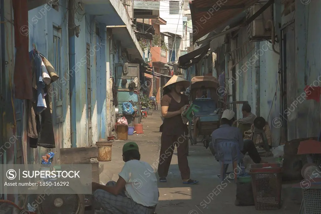 Vietnam, South, Ho Chi Minh City, View Down A Side Street With A Woman Wearing A Conical Hat And People Sat On The Ground