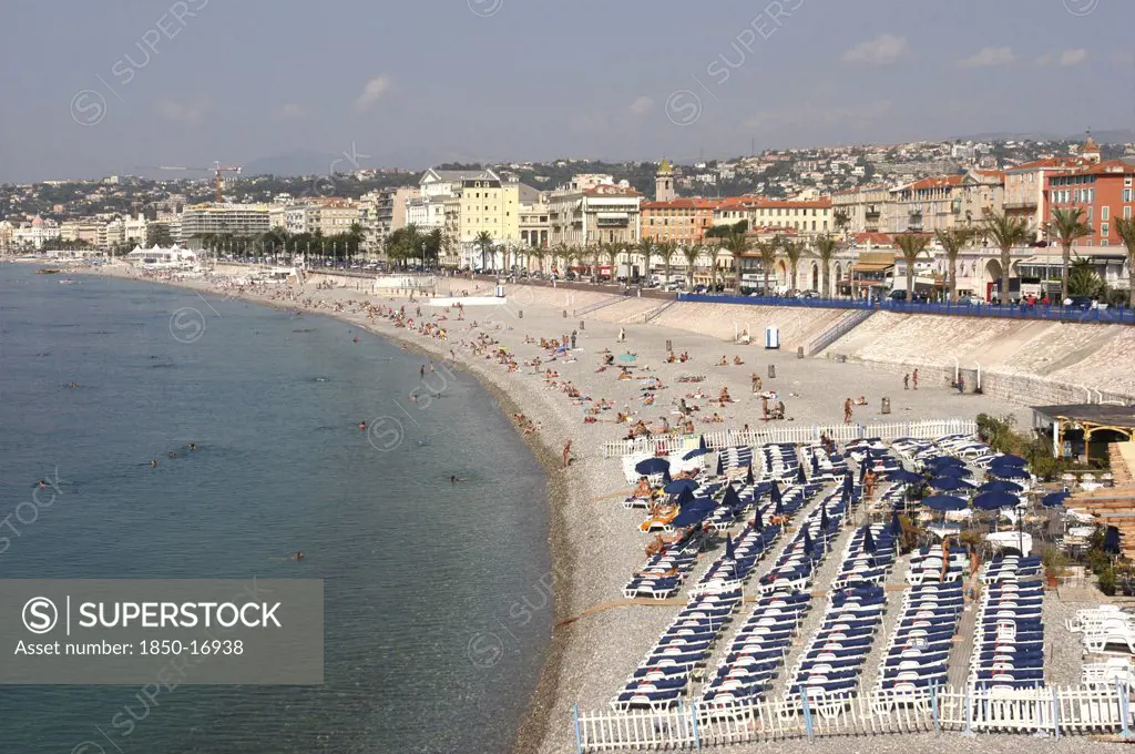 France, Nice, 'View Along The Pebbly Beach, Rows Of Sun Loungers, People Swimming And Sun Bathing. Palm Trees Line The Sea Front With Shops And Buildings.'
