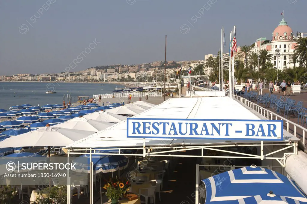 France, Nice, 'View Over Restaurant And Bar Sign Along The Pebbly Beach, Tables With Parasols Under Canopy, Blue Chairs On Promenade.'