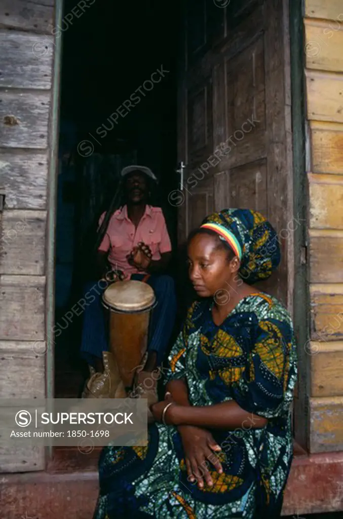 Jamaica, Music, Percussion, Rastafarian Couple With Man Playing Bongo Drums Inside Home While Woman Sits In Open Doorway.