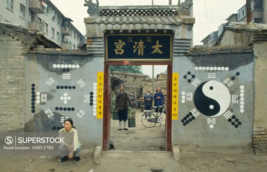 China, Inner Mongolia, Hohhot, Monks Or Priests Seen Through The Entrance Of Daoist Temple With Trigram And Yin Yang Symbols On The Exterior Walls And Person Crouched Outside.