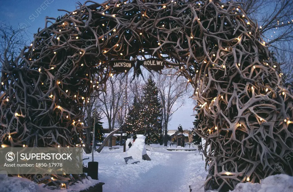 Usa, Wyoming, Jackson Town Square. View Through Elk Horn Archway Decorated With Lights Towards Snow And A Christmas Tree.