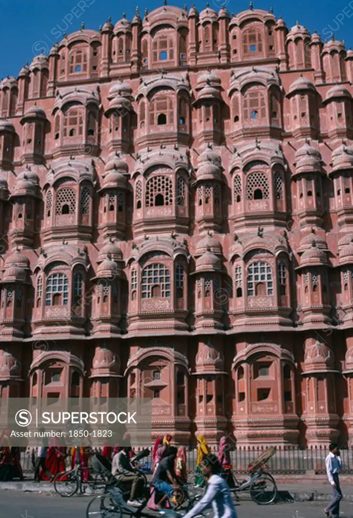 India, Rajasthan, Jaipur, 'Hawa Mahal Or Palace Of The Winds.  Part View Of Semi-Octagonal, Honeycombed Sandstone Facade.   Cyclists And Brightly Dressed Women On Street In Front. '