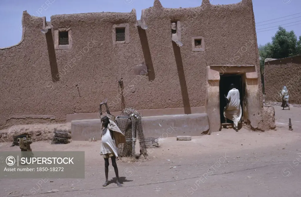 Nigeria, Kano, Traditional Mud Hausa Dwelling With Child Standing With Up-Stretched Arms Outside And Man Entering Through Open Doorway.
