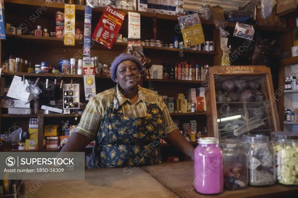 South Africa, Gauteng, Soweto, Portrait Of Shopkeeper Behind Counter With Goods Arranged On Shelves Behind Her.