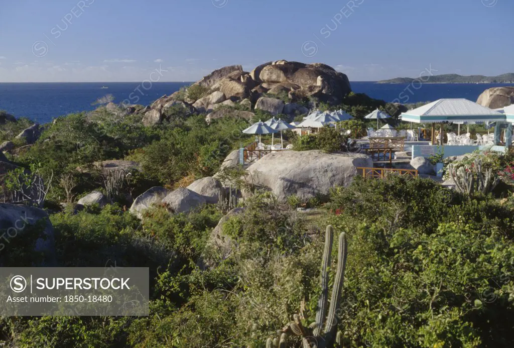 British Virgin Islands, Virgin Gorda, The Baths. Granite Boulders Amongst Plants And Vegetation With Chairs And Sun Shades From A Restaurants Open Air Verandah In The Middle. The Ocean Seen In The Distance