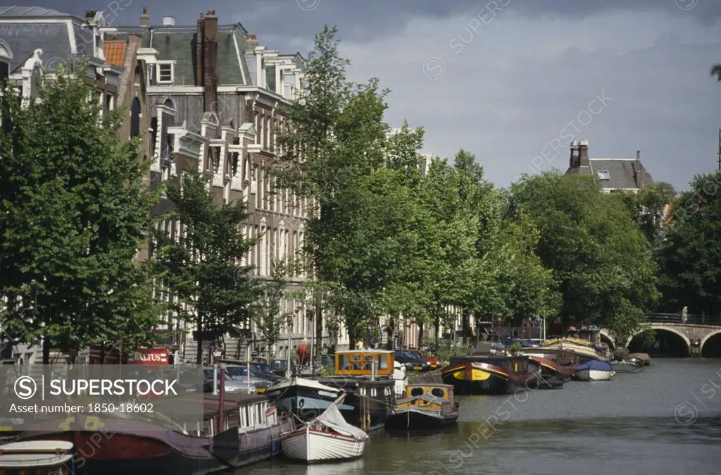 Holland, Amsterdam, Canal And Moored Houseboats And Barges Overlooked By Traditional Architecture.