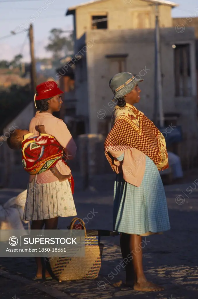 Madagascar, People, Women, Road To Fianarantsoa. Two Women One Carrying A Baby In A Sling On Her Back Wearing Colourful Clothing Standing Waiting For A Lift With Golden Sunlight Shining Onto Them