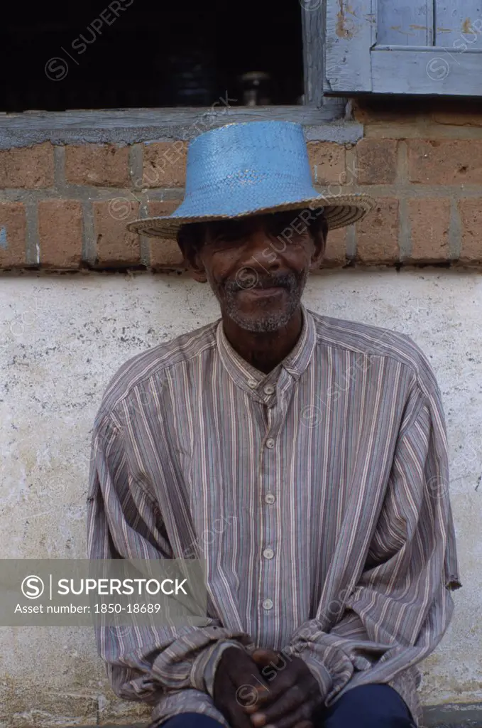 Madagascar, Ihosy, Portrait Of An Elderly Man Sitting Against A Wall Wearing A Blue Straw Hat