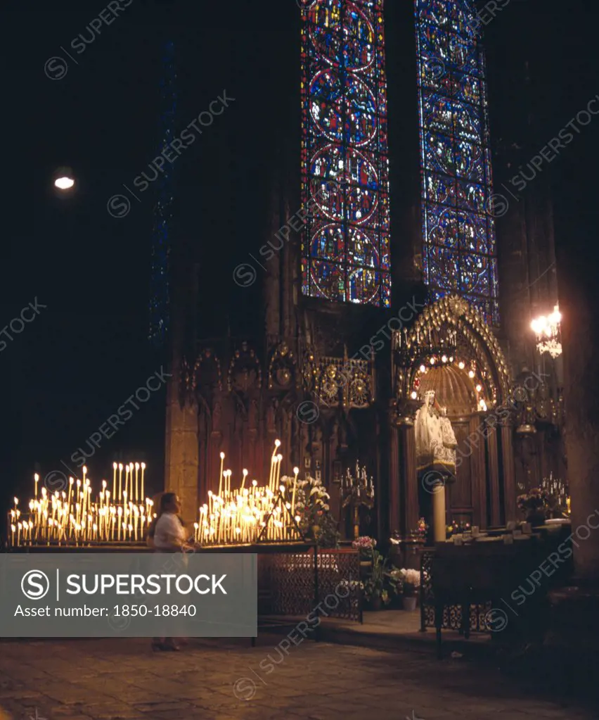 France, Central Loire, Chartres, Interior Of Cathedral Showing Notre Dame Du Pillar Black Madonna Beneath Stained Glass Window And Women Lighting Candles.