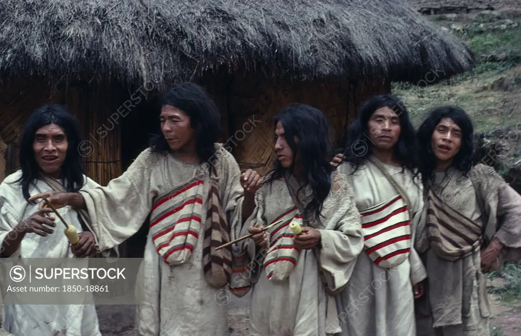 Colombia, Indigenous Tribes, Kogi, Chendukua. A Group Of Kogi Men Outside A Grass-Thatched Village Home On S.Side Of Sierra. Drunk On Aguadiente/Distilled Sugarcane Liquor -Sold To Indians By Colombian Campesinos/Peasants Hand-Woven White Cotton And Darker Fique/Cactus-Fibre Mochilas/Bags Around Necks. Two Carrying Lime Gourds And Sticks To Transfer Powdered Lime Into Wads Of Coca-Leaves In Their Mouths - Lime Acts As Catalyst To Release Very Small Amount Of Cocaine Alkaloid.
