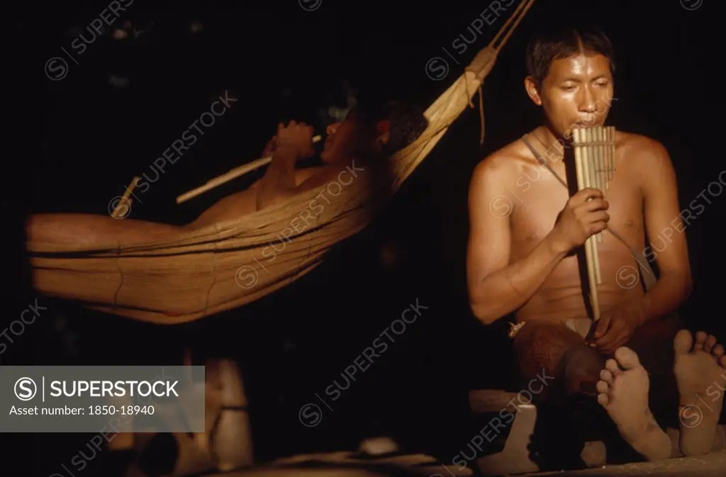 Colombia, Vaupes Region, Tukano Tribe, 'Boys Playing Panpipes At Dusk In Entrance To Maloca / Longhouse. One Sits On An Elders Stool, Other Lies In A Cumare / Fibre Hammock'