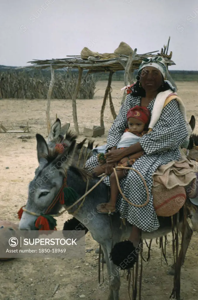 Colombia, Guajira Peninsula, Guajiro / Wayuu Tribe, 'Portrait Of Wayuu Mother With Baby Daughter. Facial Protection Against Strong Sun And Wind From Mixture Of Burnt Fungi And Goat Fat. Mother Wears Woollen Pom-Poms On Feet To Protect Toes And As Adornment, Donkey Also Decorated With Woollen Pom-Poms.'