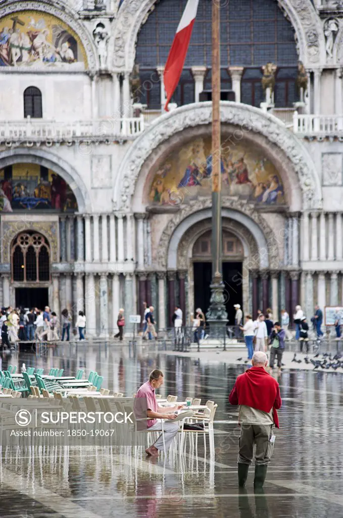 Italy, Veneto, Venice, Aqua Alta High Water Flooding In St Marks Square Showing St Marks Basilica At The End Of The Flooded Piazza With An Artist Painting A Watercolour Seated At A Table In The Water
