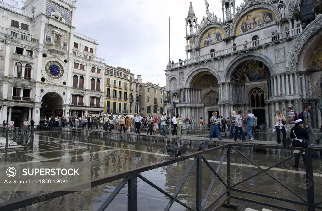 Italy, Veneto, Venice, Aqua Alta High Water Flooding In St Marks Square With Tourists On Elevated Walkways Above The Flooded Piazza Beside St Marks Basilica And The Torre Dell'Orologio Clock Tower