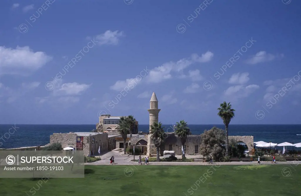 Israel, Caesarea, The Herodian Harbour With Eastern View Of Stone Buildings With Palm Trees And Visitors Walking Past Along Path.