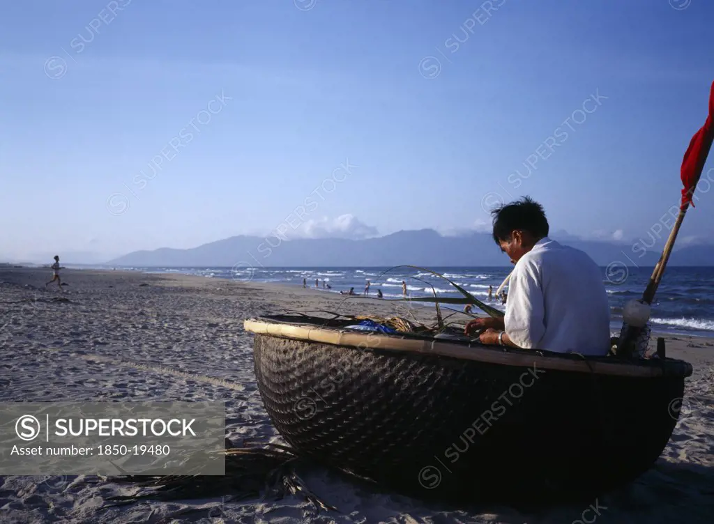 Vietnam, Central, China Beach, Fisherman Mending Nets In Coracle Boat Pulled Up Onto Sandy Beach.