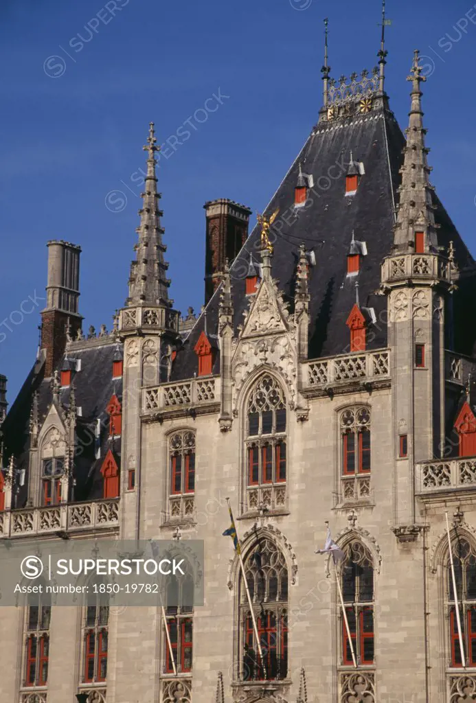 Belgium, West Flanders, Bruges, The Markt (Market Place). Exterior Facade Of Old Town Building With Red Painted Window Frames  Roof Vents And Gables With Spires And Gold Statue Of The Archangel Michael Slaying The Devil.