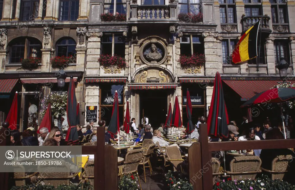 Belgium, Brabant, Brussels, Grand Place.  Busy Cafe With People Sitting At Outside Tables In The Sunshine.  Part View Of Building Facade With Stone Balcony And Flower Filled Window Boxes. Unesco World Heritage Site