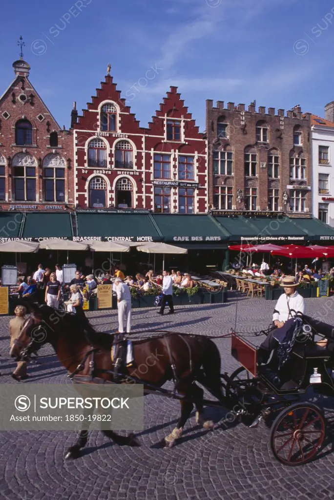Belgium, West Flanders, Bruges, The Markt (Market Place).  Horse Drawn Carriage Passing Line Of Cafes With Busy Outside Tables Under Green Awnings And Red Umbrellas.