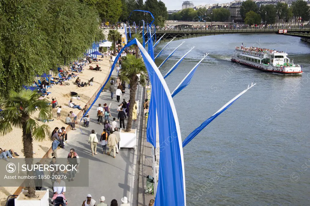 France, Ile De France, Paris, The Paris Plage Urban Beach. People Strolling Between The River Seine And Other People Lying On Sand Along The Voie Georges Pompidou Usually A Busy Road Now Closed To Traffic. A Pleasure Boat On The River With Tourists Passes Under The Pont D'Arcole Bridge
