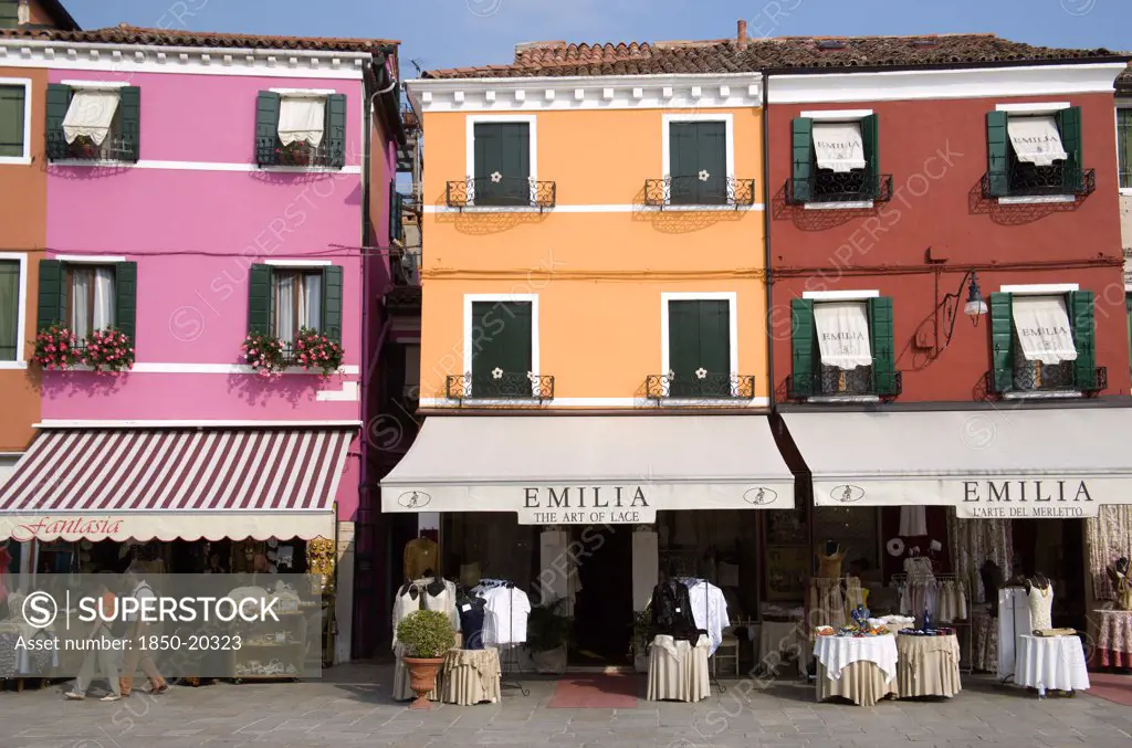 Italy, Veneto, Venice, Brightly Coloured Houses Above Lace Shops On The Lagoon Island Of Burano The Historic Home Of The Lace Making Industry In The Region. Tourists Walk Past Looking At The Displays On The Pavement