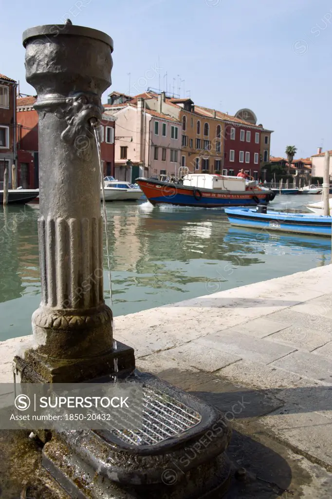 Italy, Veneto, Venice, A Free Public Drinking Water Fountain On The Canale Di San Donato On The Island Of Murano. One Of Many On The Island. A Lagoon Barge Passes Down The Canal