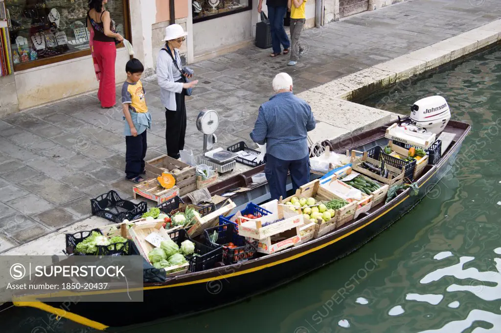 Italy, Veneto, Venice, A Fruit And Vegetable Vendor Serving A Customer From His Boat Moored Alongside The Fondamenta Dei Vetrai Along The Rio Dei Vetrai Canal On The Lagoon Island Of Murano