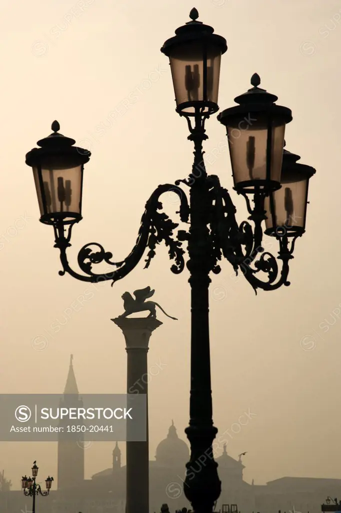 Italy, Veneto, Venice, An Ornate Lamp Post And The Column Of Saint Mark In The Piazzetta With Palladio'S Church Of San Giorgio Maggiore On The Island Of The Same Name In The Distance On A Misty Day