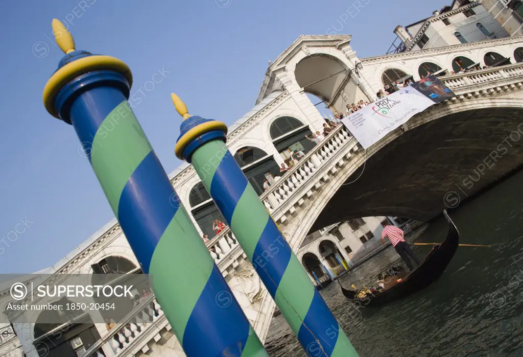 Italy, Veneto, Venice, Colourful Posts For Mooring Boats In Front Of Gondolas Carrying Sightseers On The Grand Canal Beneath The Rialto Bridge Lined With Tourists