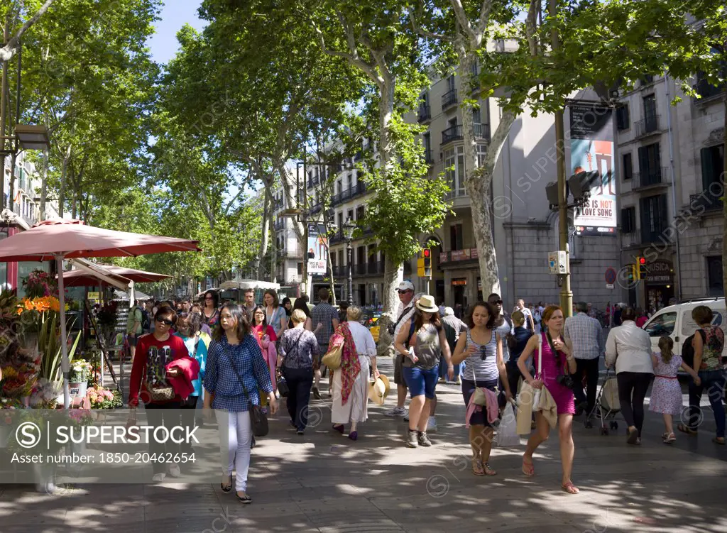 Spain, Catalonia, Barcelona, People walking along the central tree shaded walkway of La Rambla historic avenue past flower stalls in the Old Town district.
