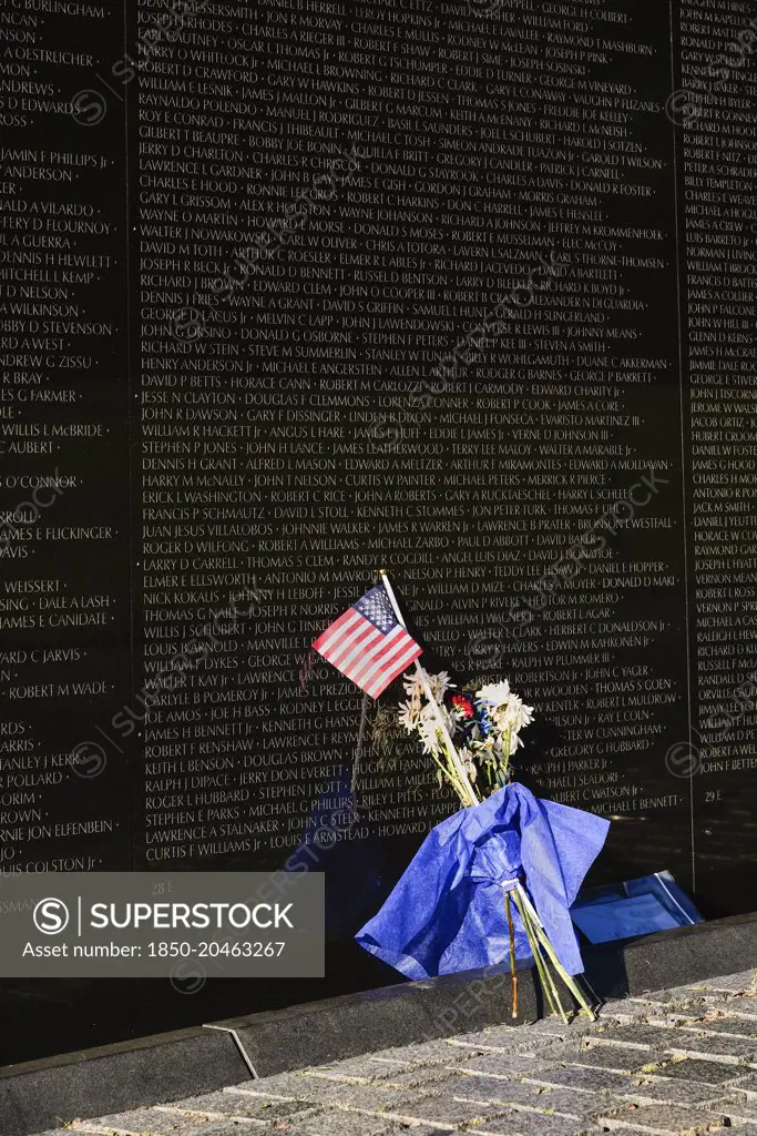 USA, Washington DC, National Mall, Vietnam Veterans Memorial, A section of The Memorial Wall with the names of those killed or missing in action during the Vietnam War, Bunch of flowers and American flag resting against the wall.
